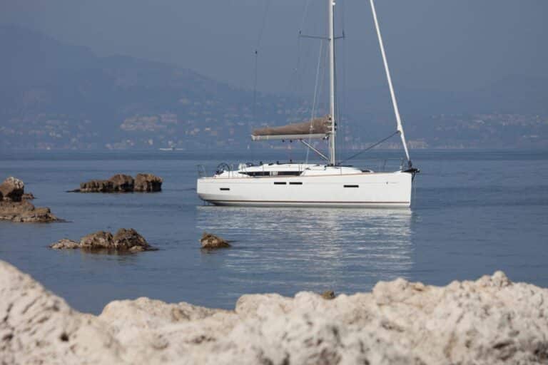 Eine weiße Segelyacht ankert in Küstennähe in ruhigen Gewässern, mit einem felsigen Vordergrund und dunstigen Hügeln im Hintergrund.