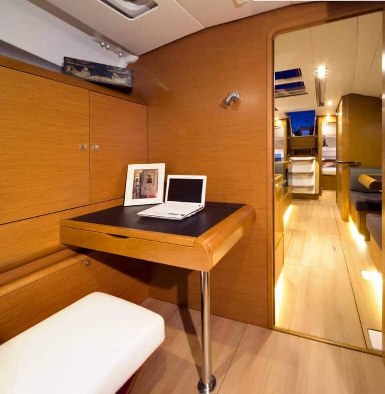 Innenansicht einer luxuriösen Segelyacht mit kleinem Holzschreibtisch und Laptop, weißem Hocker und geschwungenen Holzwänden, die im Hintergrund zu mehr Kabinenraum führen.