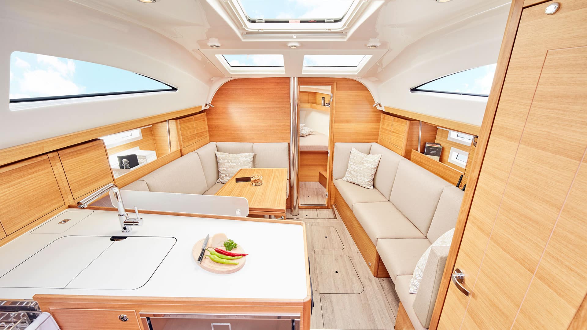 Innenansicht einer modernen Segelyachtkabine mit hellen Holzpaneelen, weiß gepolsterten Sitzen, einem zentralen Tisch mit einer Obstschale und Oberlichtern, die für natürliches Licht sorgen.