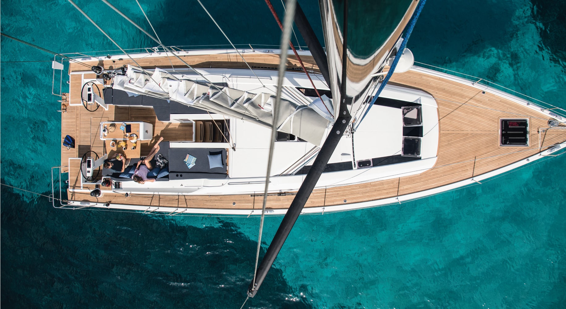 Luftaufnahme einer luxuriösen Segelyacht auf klarem, blauem Wasser. Mehrere Personen faulenzen und unterhalten sich auf dem Deck.