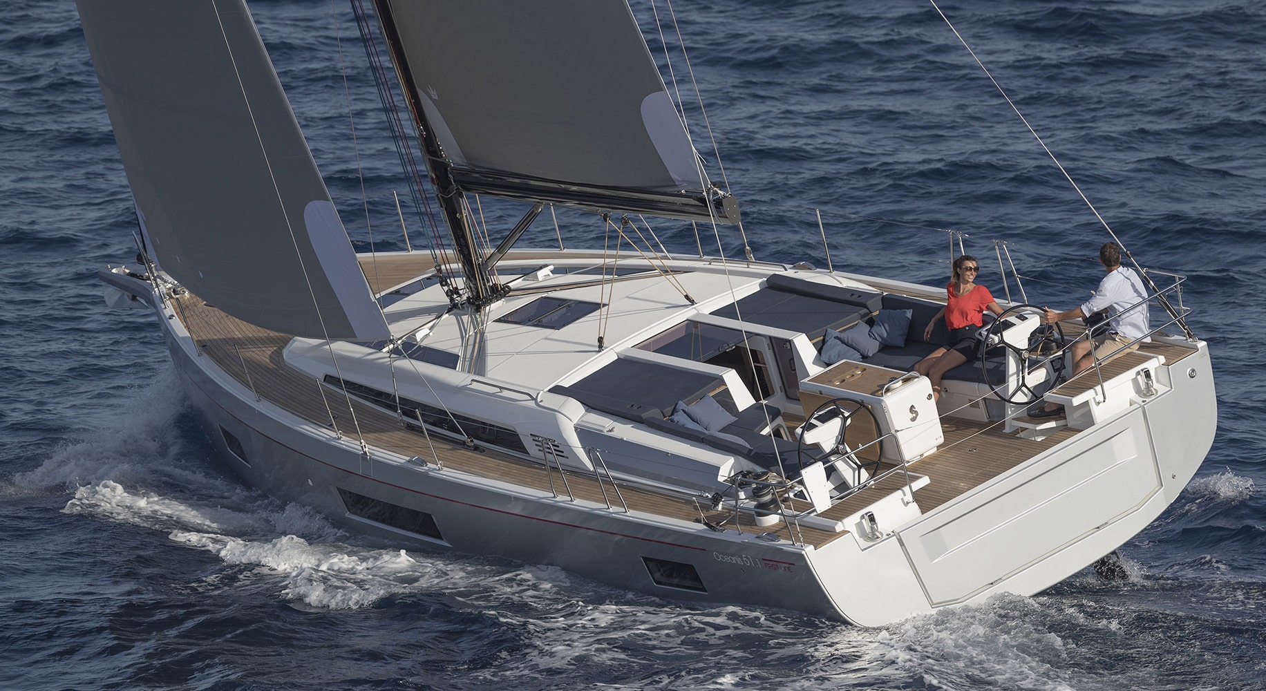 Luftaufnahme von zwei Personen auf einer Segelreise, die auf einer modernen Yacht mit weißen Segeln durch das blaue Meer segeln. Eine Person steht am Steuer, während die andere daneben sitzt.