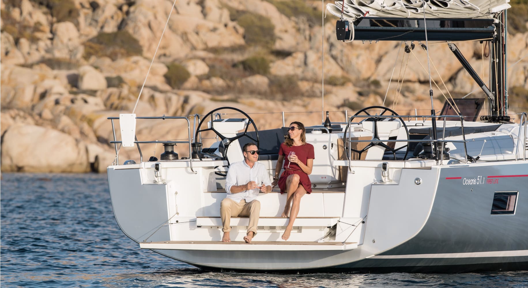 Ein Paar genießt einen ruhigen Moment auf dem Heck einer Segelyacht, die sanft an felsigen Ufern vorbeitreibt. Der Mann hält ein Weinglas und die Frau ein Getränk, beide wirken zufrieden.