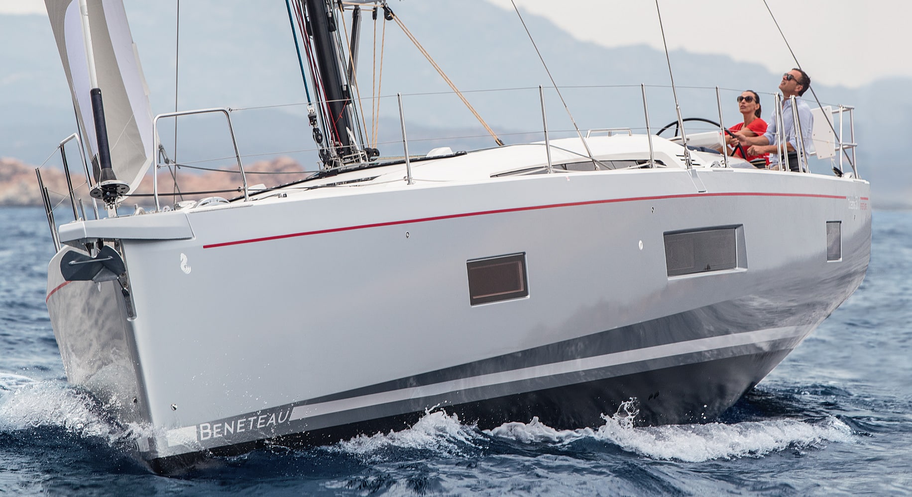 Eine elegante, moderne Segelyacht namens Beneteau segelt auf offener See. Das Boot schneidet durch die Wellen mit zwei Personen an Deck, einer am Steuer und der andere daneben. Im Hintergrund sind Berge und ein klarer Himmel zu sehen, die für einen unvergesslichen Segeltörn sorgen.