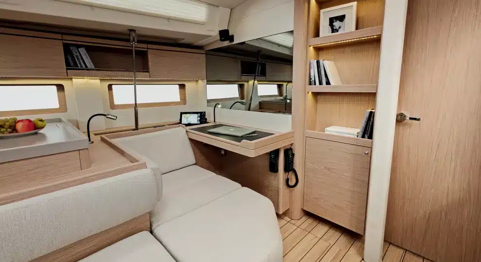 Innenansicht einer modernen Segelyacht mit einem gemütlichen Wohnbereich mit heller Holzvertäfelung, einem geschwungenen Sofa, einem kompakten Küchenbereich und mehreren Fenstern für natürliches Licht.