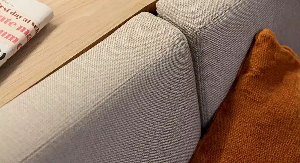 Nahaufnahme einer hellgrau strukturierten Sofaecke mit einem rostorangen Samtkissen und einer unscharfen Zeitung im Hintergrund, die eine gemütliche häusliche Atmosphäre während einer Segelreise suggeriert.