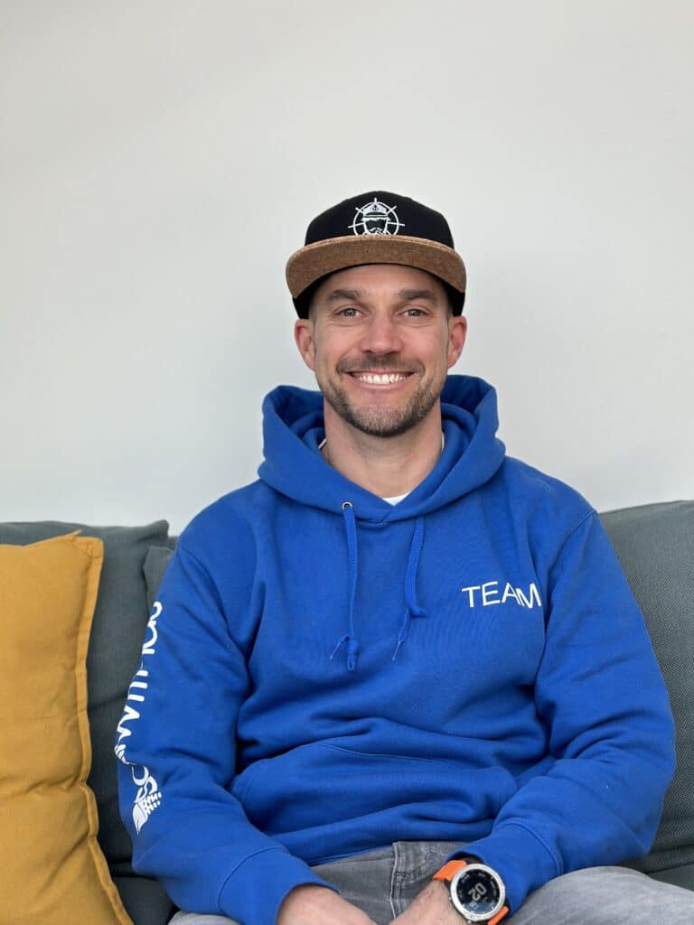 Ein Mann sitzt auf einem Sofa. Er trägt einen blauen Kapuzenpullover mit „TEAM“-Aufdruck auf der Brust und eine schwarze Kappe mit einer weißen Grafik auf der Vorderseite. Er trägt eine Armbanduhr am linken Handgelenk und lächelt. Neben ihm ist ein gelbes Kissen zu sehen, das an Entspannung nach einem abenteuerlichen Segelurlaub erinnert. Der Hintergrund ist schlicht weiß.