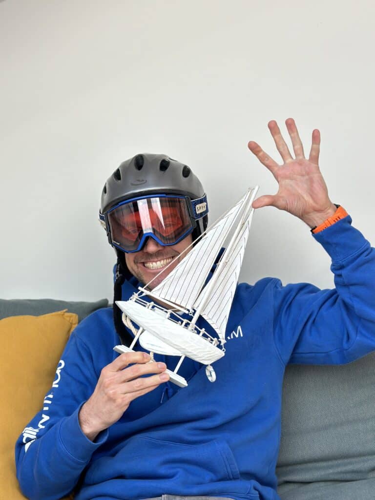 Eine Person mit schwarzem Helm und Skibrille sitzt auf einem Sofa, lächelt und winkt mit einer Hand, während sie mit der anderen ein kleines weißes Modellsegelboot hält. Sie trägt einen blauen Kapuzenpullover und scheint bereit für ihr nächstes Segelurlaubsabenteuer. Ein gelbes Kissen neben ihr sorgt für Bequemlichkeit.