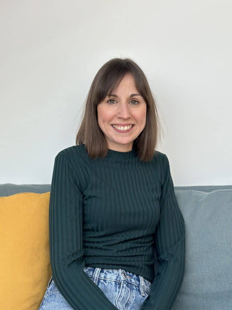 Eine Frau mit schulterlangem braunem Haar und einem grün gestreiften Langarmshirt sitzt auf einem Sofa, neben ihr liegt ein gelbes Kissen. Sie lächelt und blickt in die Kamera. Sie strahlt Wärme und Freundlichkeit aus, was an eine unterstützende Teamumgebung erinnert. Der Hintergrund ist eine schlichte weiße Wand.