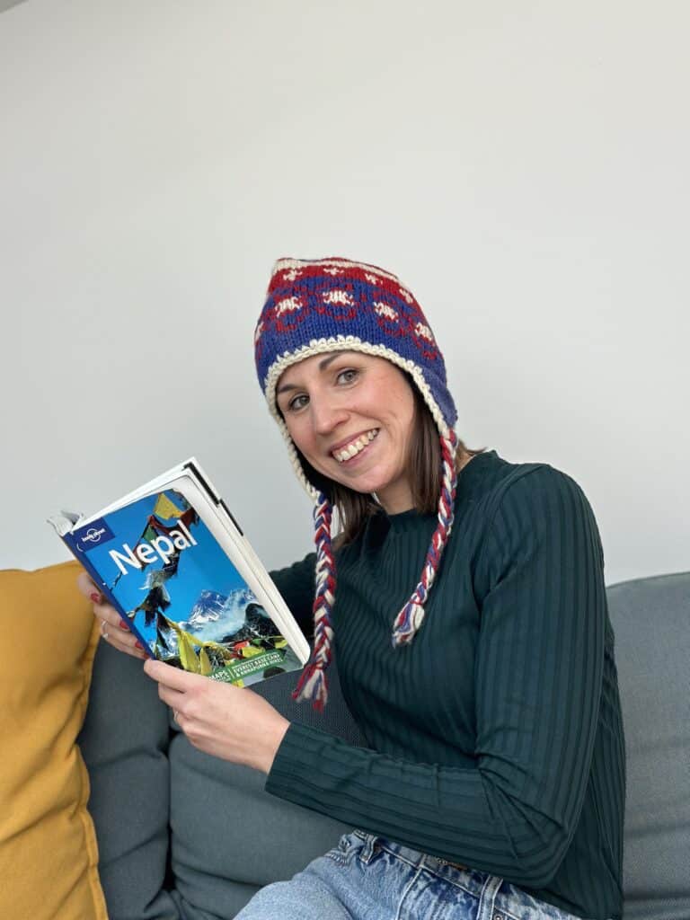 Eine Frau mit einer bunten Strickmütze mit Ohrenklappen und einem grünen Pullover sitzt lächelnd auf einem grauen Sofa. Sie hält einen Reiseführer mit dem Titel „Nepal“ in der Hand und sieht ihn sich an. Vielleicht träumt sie von ihrem nächsten Abenteuer, sei es eine Wanderung im Himalaya oder die Planung eines Segeltörns. Neben ihr auf dem Sofa liegt ein gelbes Kissen.