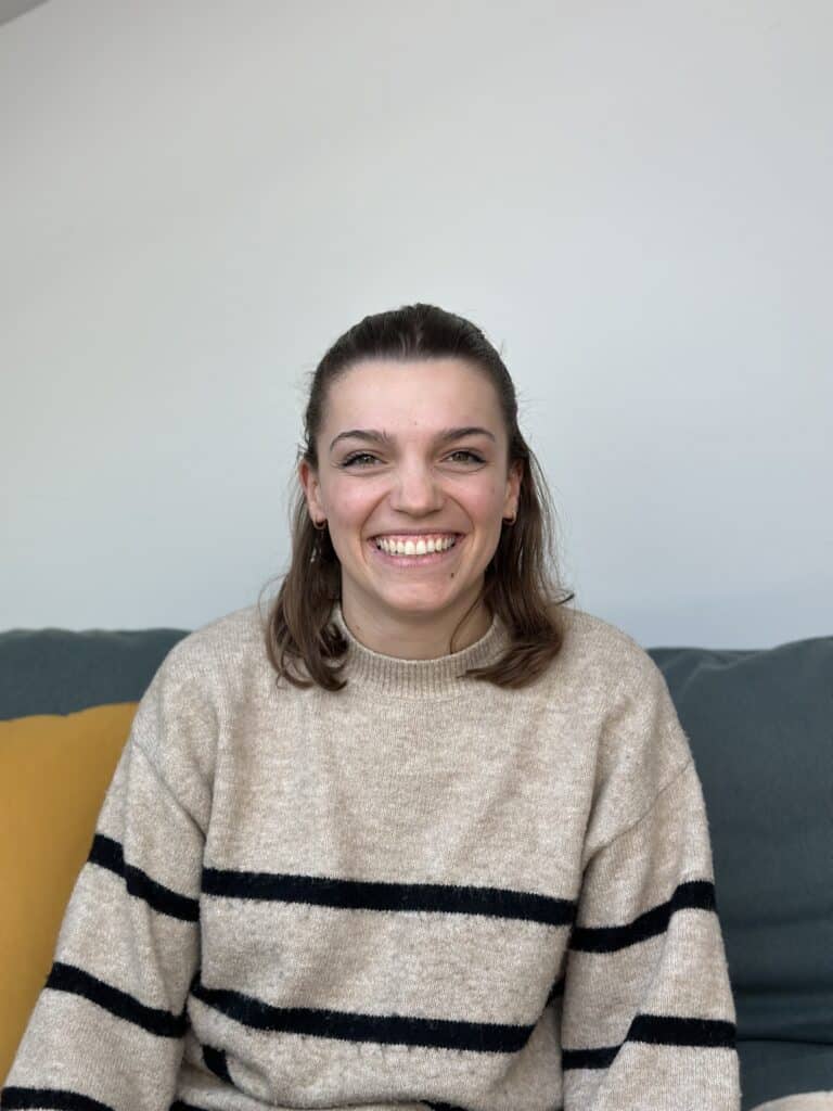 Eine Person mit schulterlangem braunem Haar sitzt auf einem Sofa und lächelt in die Kamera. Sie trägt einen beigen Pullover mit schwarzen Querstreifen. Im Hintergrund ist eine helle Wand zu sehen, auf der linken Seite ist ein Teil eines gelben Kissens zu sehen, was gemütliche Erinnerungen an ihr jüngstes Segeltörn-Abenteuer weckt.