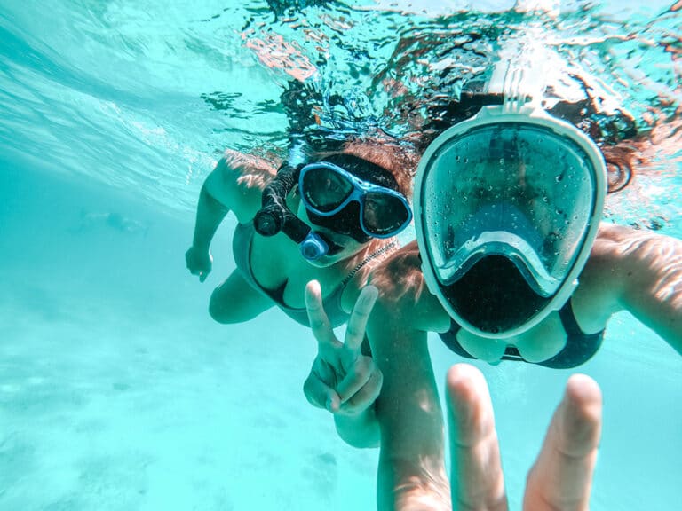 Zwei Personen unter Wasser mit Schnorchelmasken machen während ihres Segelurlaubs ein Selfie. Eine Person hält mit den Fingern ein Peace-Zeichen hoch. Sie sind umgeben von klarem blauem Wasser in der Nähe ihres Katamarans.