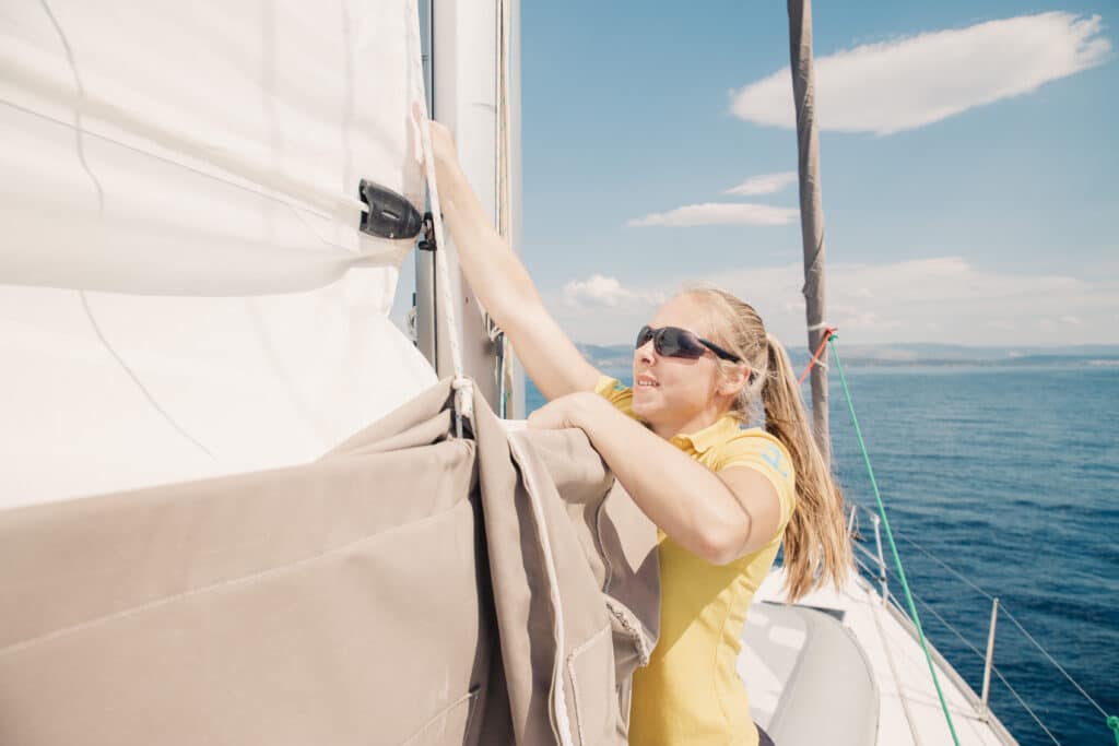 Eine Person mit Sonnenbrille und gelbem Hemd justiert das Segel einer Segelyacht unter einem klaren, sonnigen Himmel. Das Boot liegt auf ruhigem, blauem Wasser, im Hintergrund ist eine entfernte Küstenlinie zu sehen. Die Person wirkt konzentriert, während sie während ihrer Segelreise das Segel bedient.