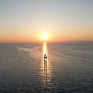 Ein Katamaran gleitet bei Sonnenuntergang auf ruhiger See dem Horizont entgegen. Die goldene Sonne wirft einen schimmernden Lichtstreifen auf die Wasseroberfläche. In der Ferne sind unter einem klaren, von Orange nach Blau wechselnden Himmel schwach kleine Inseln zu erkennen, was dieser Segelreise einen malerischen und ruhigen Charakter verleiht.