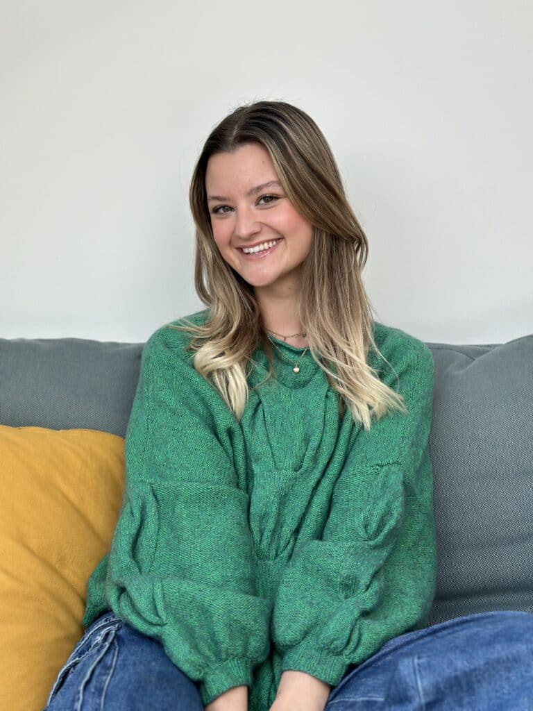 Eine Frau mit langen blonden Haaren sitzt auf einem grauen Sofa mit einem gelben Kissen. Sie trägt einen weiten grünen Pullover und blaue Jeans. Die Frau, die gerade von einer aufregenden Segelreise zurückgekehrt ist, lächelt herzlich vor dem schlichten weißen Wandhintergrund.