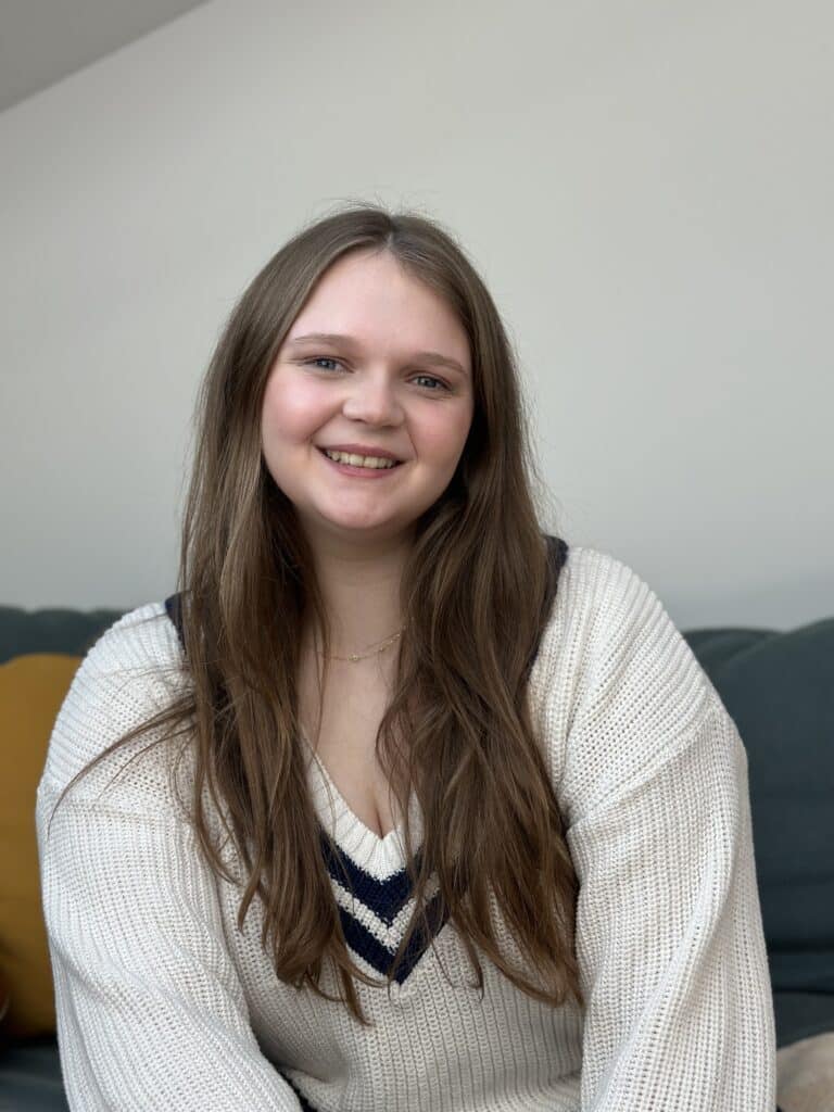 Eine junge Frau mit langen braunen Haaren lächelt in die Kamera. Sie trägt einen weißen Pullover mit marineblauem und schwarzem Ausschnitt, der an ihre jüngste Segelreise erinnert. Im Hintergrund sind ein graues Sofa und ein senffarbenes Kissen zu sehen.