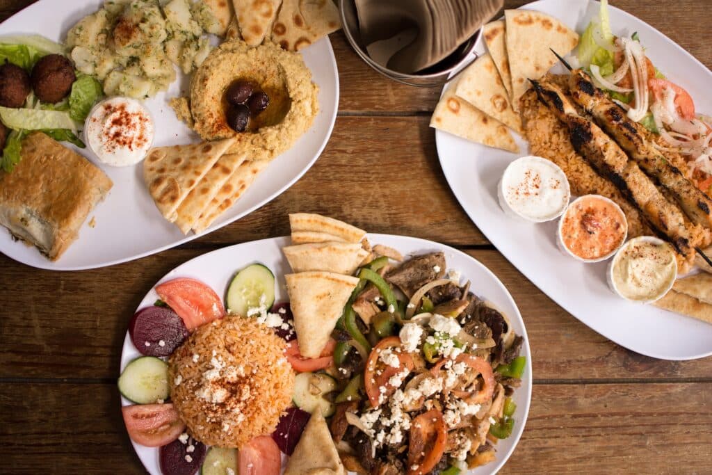 Drei Teller mit mediterraner Küche auf einem Holztisch erwecken das Gefühl einer herrlichen Segelreise. Die Gerichte umfassen Pita-Brot, Hummus, Falafel, Spieße, gemischtes Grillgemüse, Salat, Reis, Tzatziki und verschiedene Dip-Saucen. Oben in der Bildmitte steht ein Korb mit gefalteten Servietten.