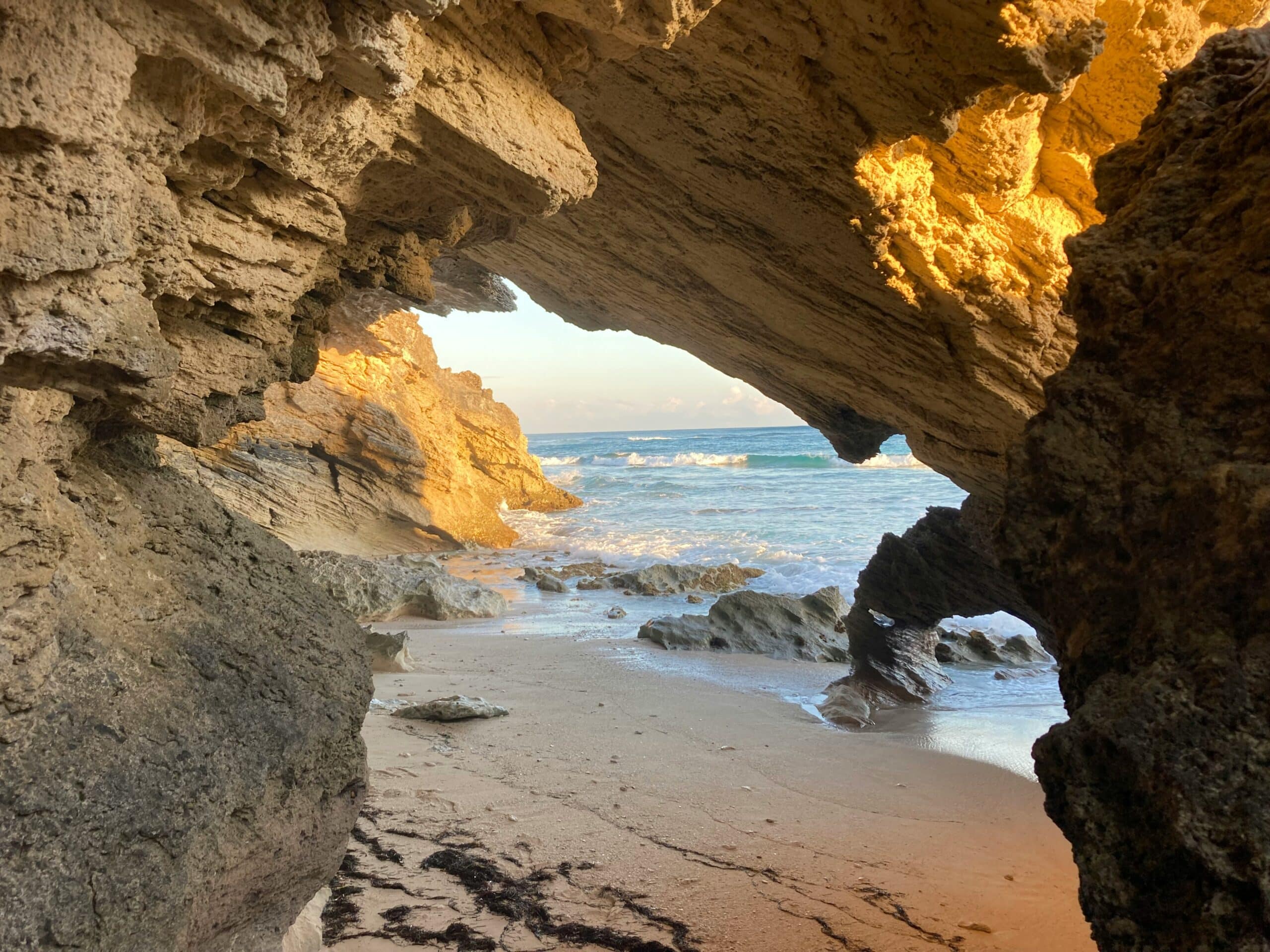 Die Öffnung einer felsigen Küstenhöhle bietet einen Blick auf einen ruhigen Sandstrand, an dessen Ufer sanft Wellen schlagen. Die Szene wird von goldenem Sonnenlicht erhellt, das warme Farbtöne auf die schroffen Felsformationen wirft und einen faszinierenden natürlichen Rahmen für die Meereslandschaft schafft, perfekt, um vorbeifahrende Katamarane auf ihrem Segeltörn zu beobachten.