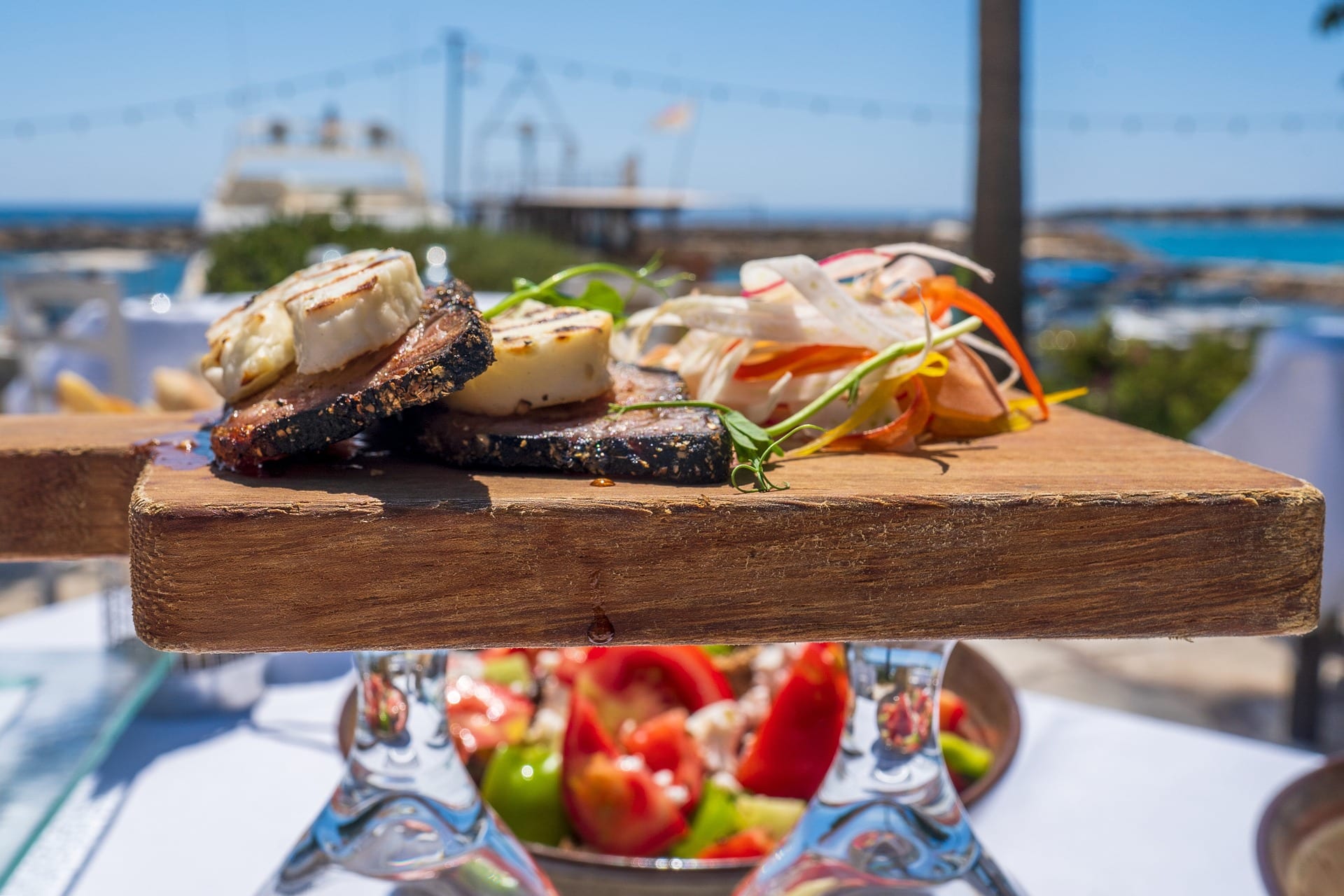 Eine Holzplatte mit gegrillten Fleischscheiben und garniertem Gemüse wird auf erhöhten Glasständern präsentiert, mit einer malerischen Außenkulisse mit Meer und blauem Himmel im Hintergrund. Im verschwommenen Hintergrund flüstert ein weiterer Teller mit buntem Gemüse die Essenz der Kykladischen Kulinarik: Entdecken Sie verborgene Schätze.