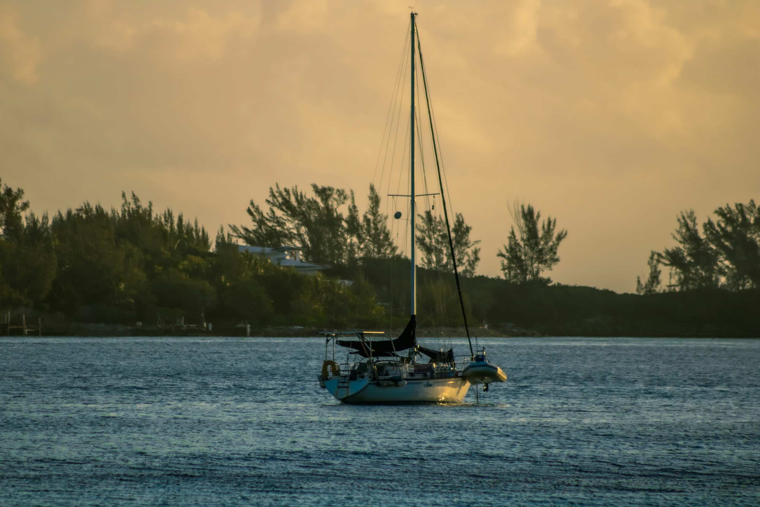Ein Segelboot schwimmt an einem ruhigen Abend sanft auf dem ruhigen Wasser in Ufernähe und verkörpert die Essenz eines heiteren Segelurlaubs. Der Himmel ist mit warmen Orange- und Gelbtönen bemalt, als die Sonne untergeht. Bäume und ein entferntes Haus zeichnen sich als Silhouetten gegen den sanft beleuchteten Himmel ab.