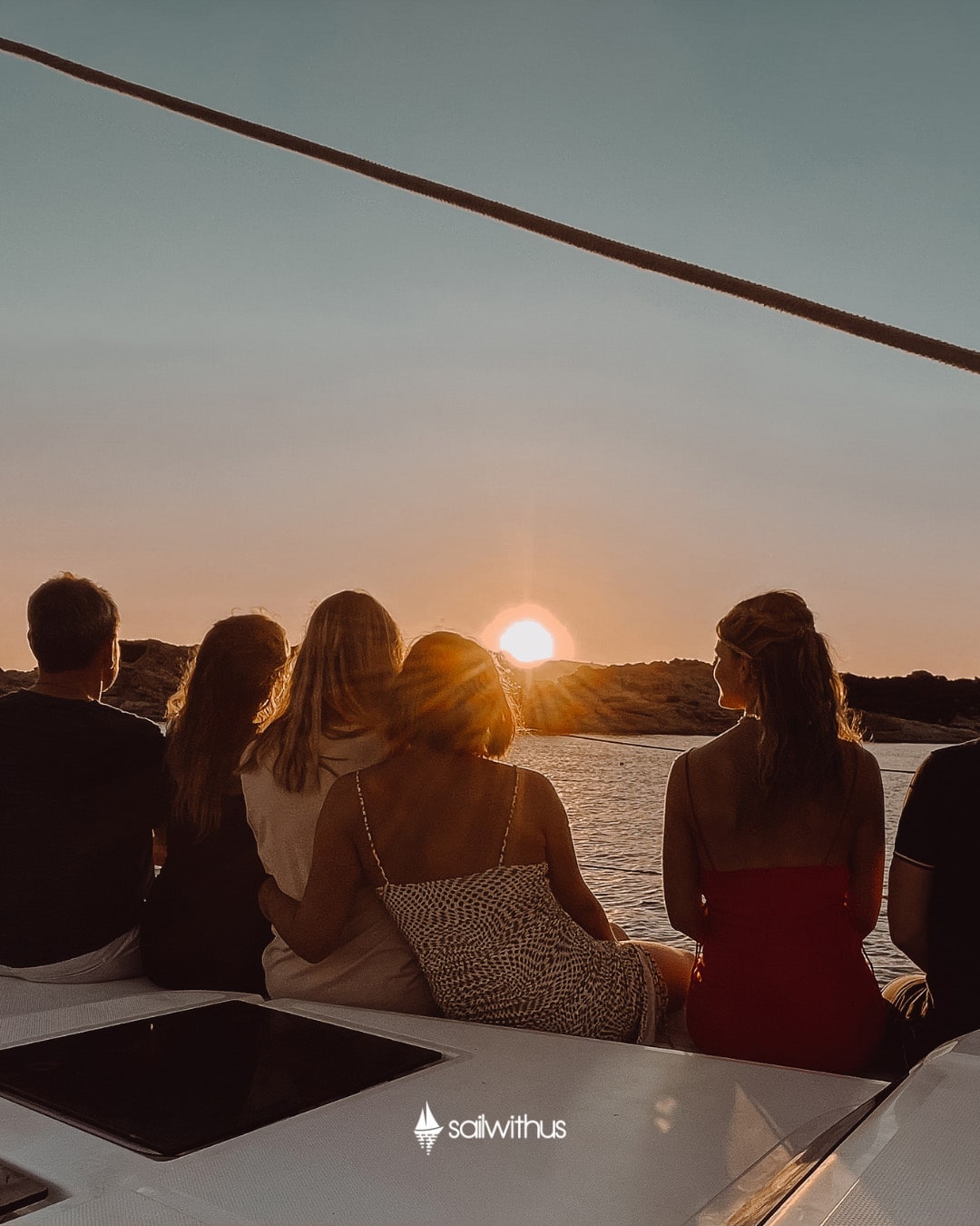 Eine Gruppe von fünf Personen sitzt auf dem Deck einer Segelyacht und beobachtet den Sonnenuntergang über dem Wasser. Sie blicken von der Kamera weg und wenden dem Betrachter den Rücken zu. Der Himmel ist in warmen Farben gehalten und die Sonne steht knapp über dem Horizont. Unten steht der Text „sailwithus“.