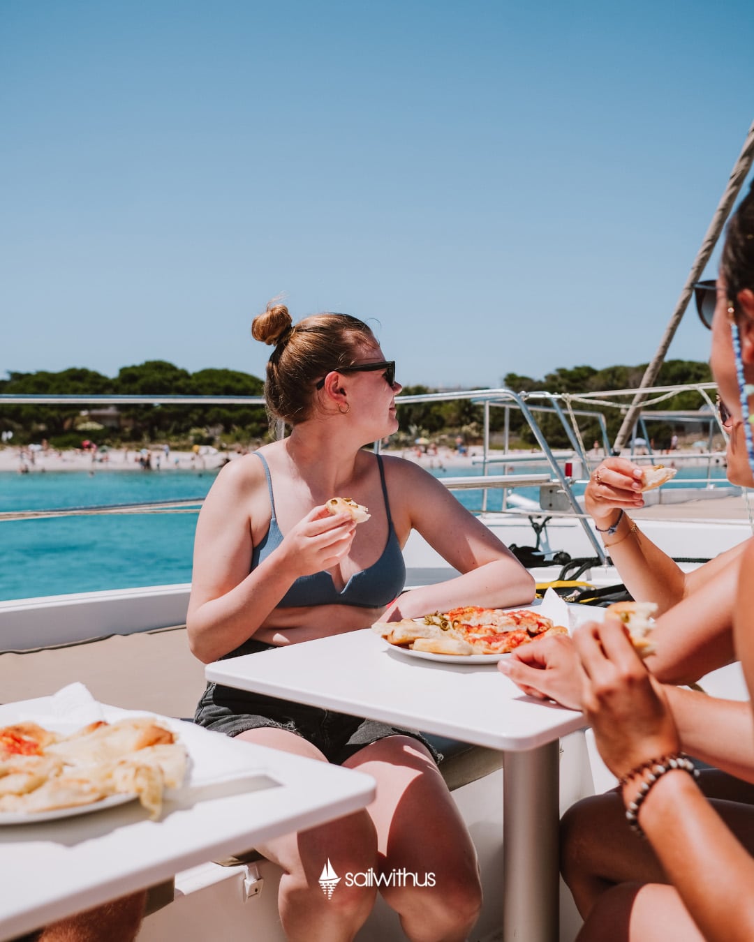 Eine Frau in Badeanzug und Shorts sitzt an einem Tisch im Freien auf einem Boot und genießt mit ihren Begleitern während ihres Segelurlaubs Pizza. Sie sind umgeben von klarem blauen Wasser und einem Sandstrand mit Bäumen im Hintergrund. Der Himmel ist klar und sonnig, was für einen perfekten Tag zum Mitsegeln sorgt.