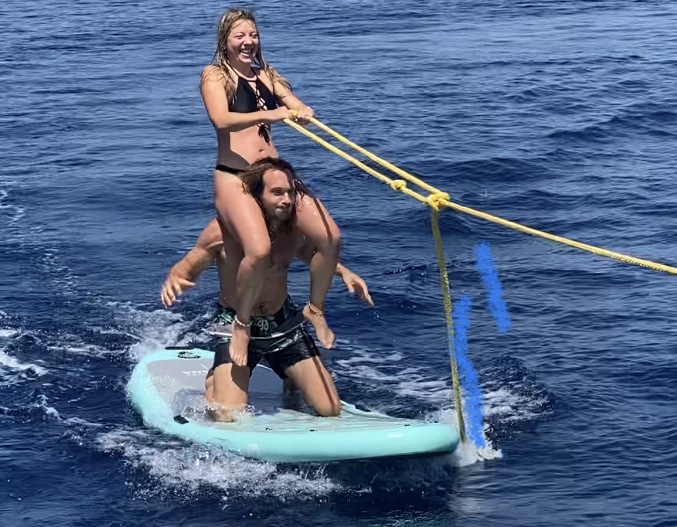 Eine Frau sitzt auf den Schultern eines Mannes, der auf einem Paddleboard im Meer kniet. Sie halten sich an einem gelben Seil fest, lächeln und genießen den sonnigen Tag, der an eine Segelreise erinnert. Der Mann balanciert auf dem hellblauen Paddleboard und steuert beide mit Leichtigkeit durch das Wasser.