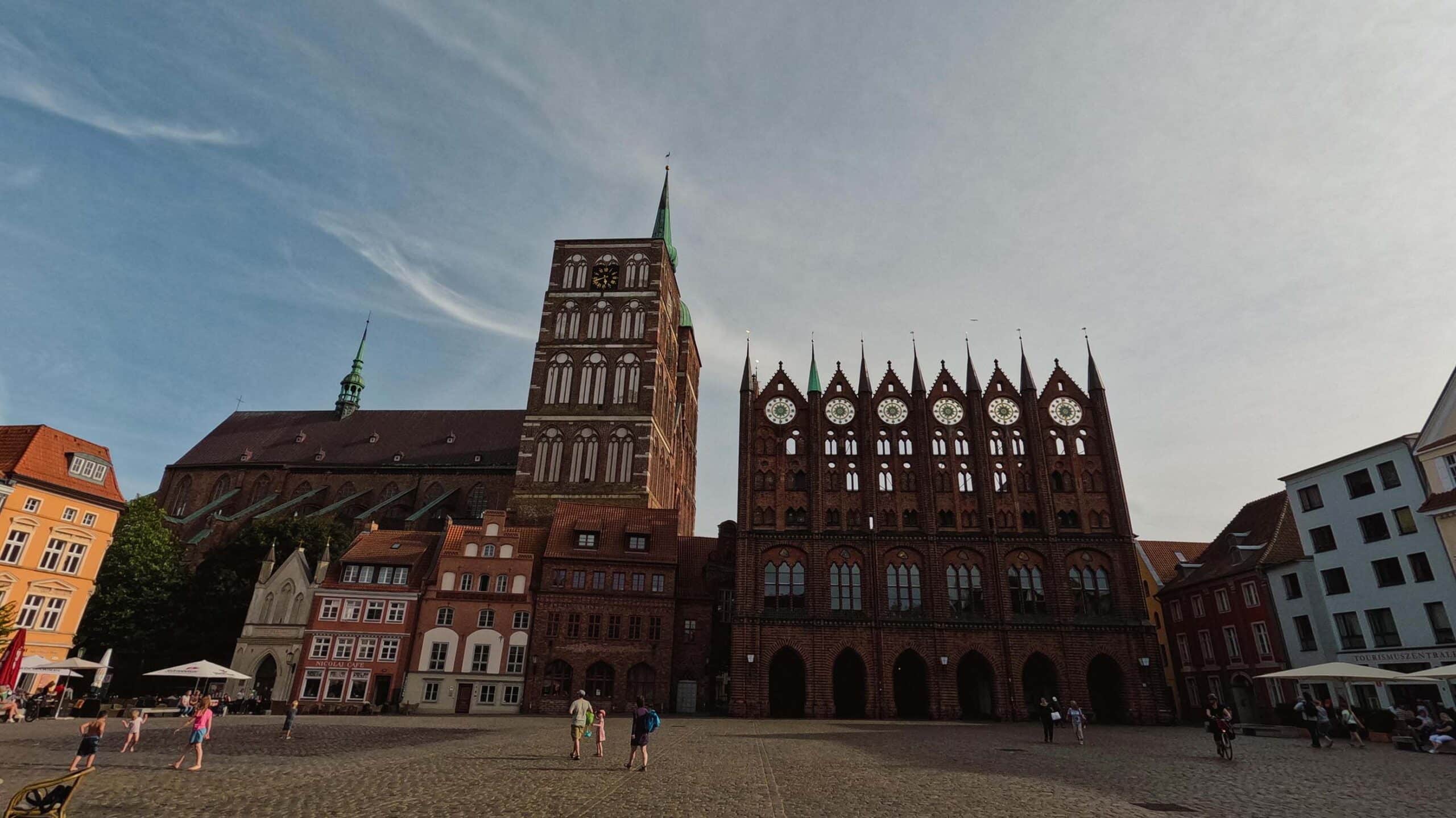 Ein historischer Stadtplatz mit einem großen, reich verzierten Gebäude im gotischen Stil mit mehreren Türmen und Zifferblättern. Um den Platz herum stehen farbenfrohe Gebäude und eine hohe Kirche mit steilem Dach und Turm. An einem sonnigen Tag gehen die Leute spazieren und sitzen in Straßencafés und erzählen sich Geschichten von ihrem letzten Segelurlaub.