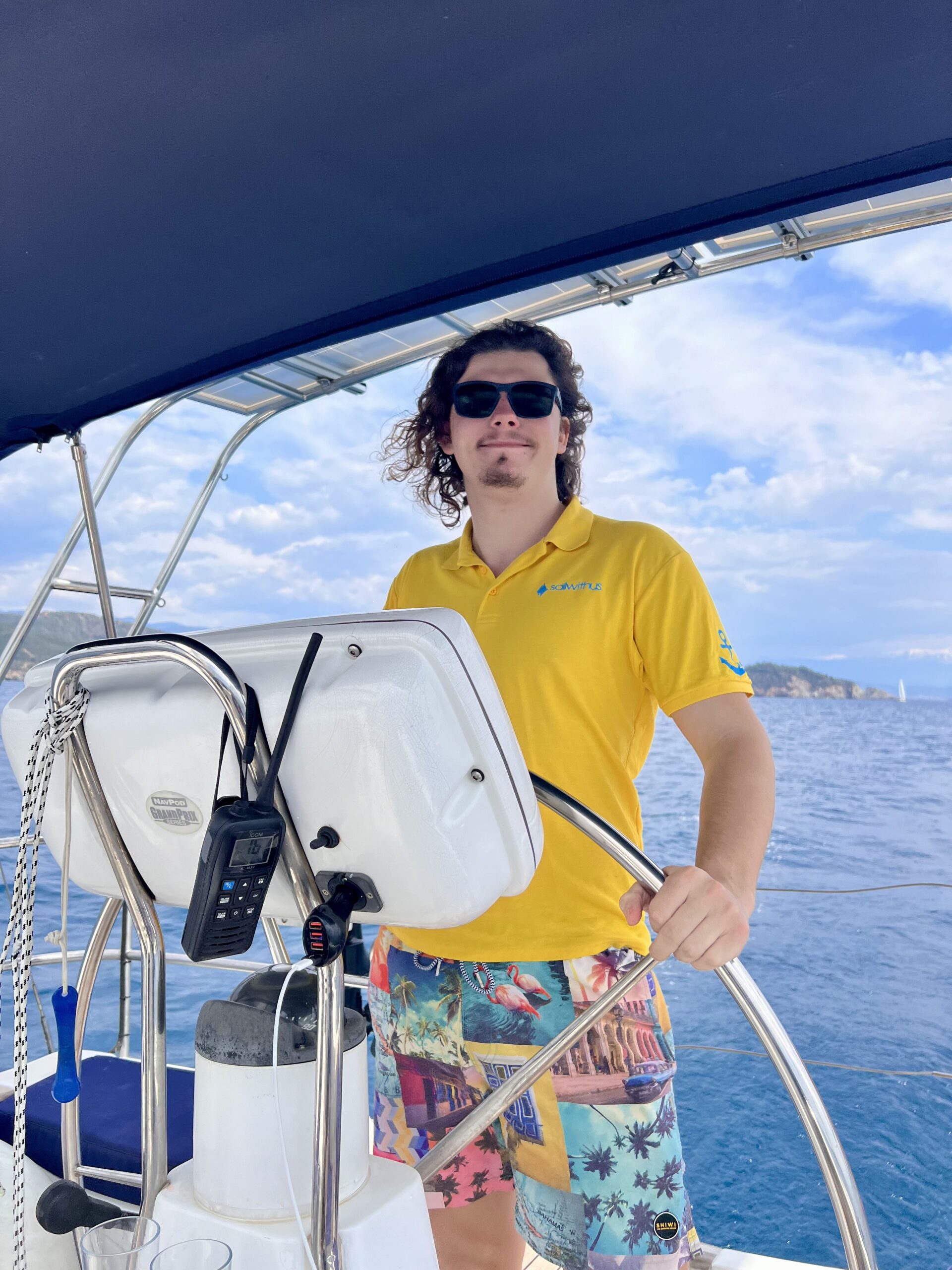 Eine Person mit langen Haaren, Sonnenbrille, gelbem Poloshirt und bunten Shorts steht während ihres Segelurlaubs am Steuer eines Bootes. Im Hintergrund sind klares blaues Wasser und eine entfernte Küstenlinie unter einem teilweise bewölkten Himmel zu sehen. In der Nähe der Person sind Fernkommunikationsgeräte zu sehen.