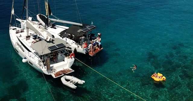 Zwei Segelboote, darunter ein schnittiger Katamaran, liegen nebeneinander im klaren blauen Wasser vor Anker. An Bord schwimmen Menschen. Eine Person schwimmt auf einem gelb-orangen Schlauchboot. Die Szene fängt einen sonnigen, entspannten Segelurlaub auf dem Wasser ein.