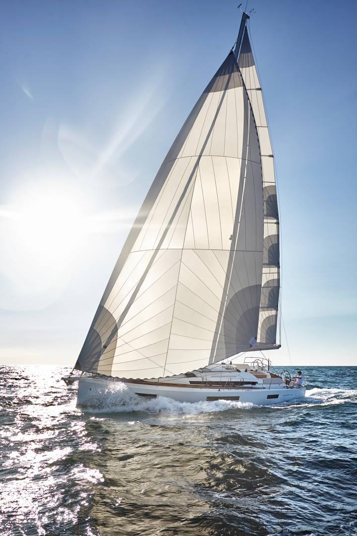 Ein Segelboot mit großen weißen Segeln gleitet unter einem strahlend blauen Himmel über das Wasser. Die Sonne scheint hell im Hintergrund, wirft einen Glanz auf das Wasser und hebt das schlanke Design des Bootes hervor. Wellen brechen sanft um das Yachtcharterboot herum, während es durch das Meer schneidet.