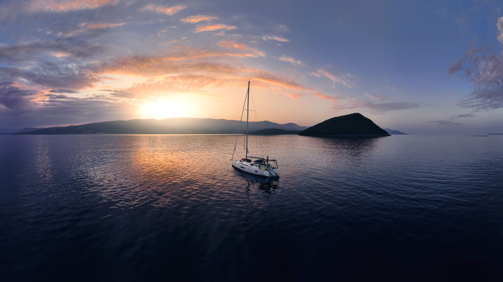 Ein Segelboot schwimmt friedlich auf einer ruhigen, weiten Wasserfläche bei Sonnenuntergang – ein perfekter Segelurlaub. Der Himmel ist erfüllt von einer dramatischen Mischung aus Farben, von tiefem Violett und Blau bis hin zu warmem Orange und Rosa, die sich im Wasser spiegeln. In der Ferne zeichnet sich eine hügelige Insel als Silhouette gegen den Horizont ab.