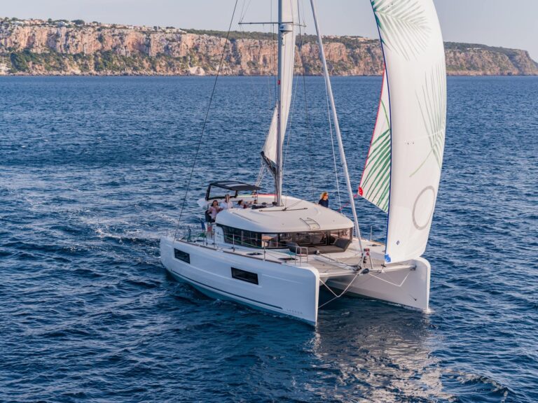 Ein weißer Youngline-Katamaran mit voll ausgefahrenen Segeln gleitet sanft über das klare, blaue Meer. Das Schiff hat mehrere Personen an Bord und nähert sich einer Küste mit hohen Klippen und spärlicher Vegetation. Im Hintergrund sind ein klarer Himmel und ferne, felsige Küsten zu sehen, die den malerischen Segeltörn vervollständigen.