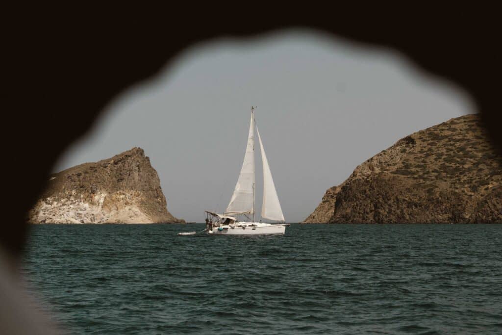 Ein weißes Segelboot gleitet auf einem ruhigen Meer, eingerahmt von felsigen Klippen auf beiden Seiten. Das Bild, das an ein Segelabenteuer erinnert, wird durch eine schlüssellochartige Öffnung betrachtet, mit dem Segelboot in der Mitte. Der Himmel ist klar und das Wasser ist tief blaugrün.