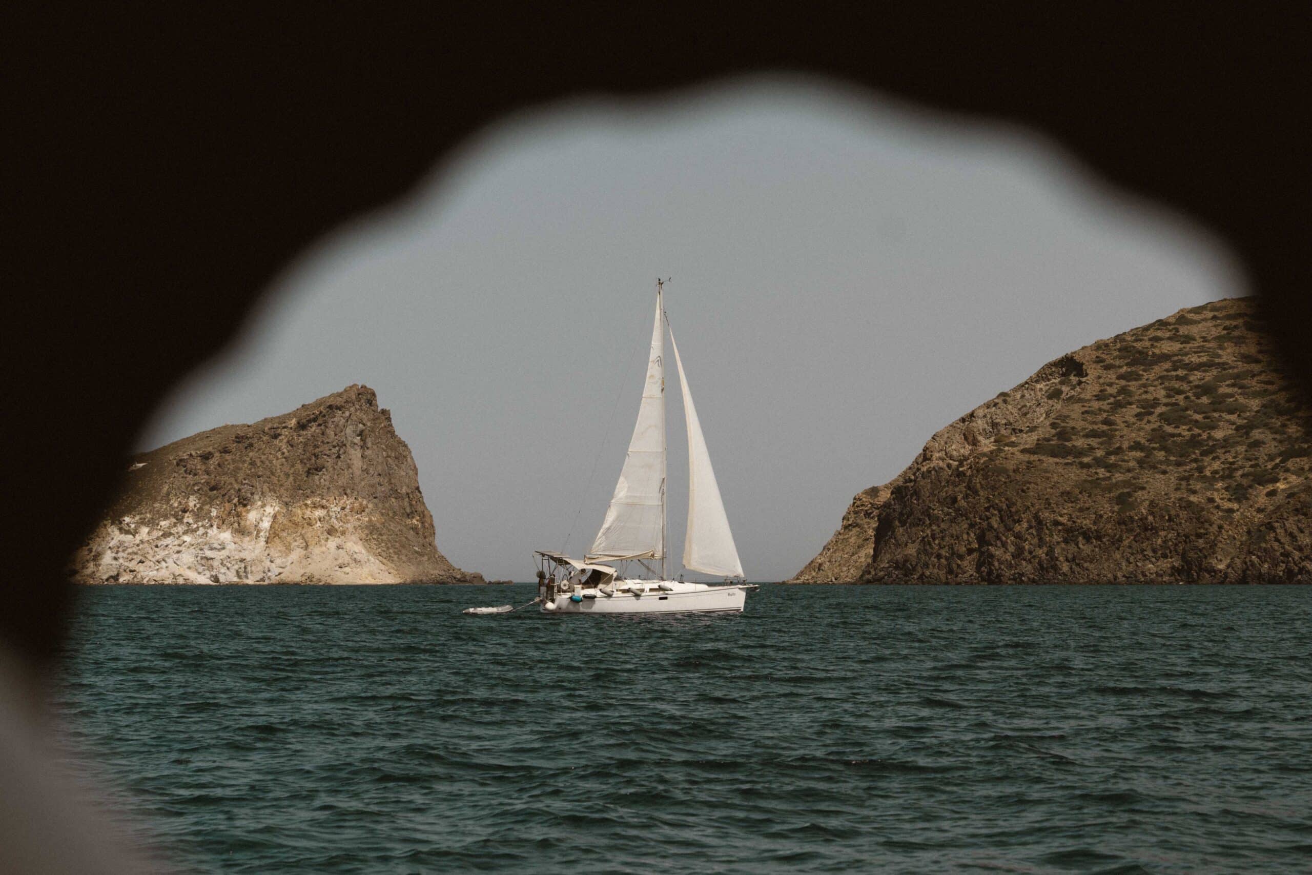 Ein weißes Segelboot gleitet auf einem ruhigen Meer, eingerahmt von felsigen Klippen auf beiden Seiten. Das Bild, das an ein Segelabenteuer erinnert, wird durch eine schlüssellochartige Öffnung betrachtet, mit dem Segelboot in der Mitte. Der Himmel ist klar und das Wasser ist tief blaugrün.