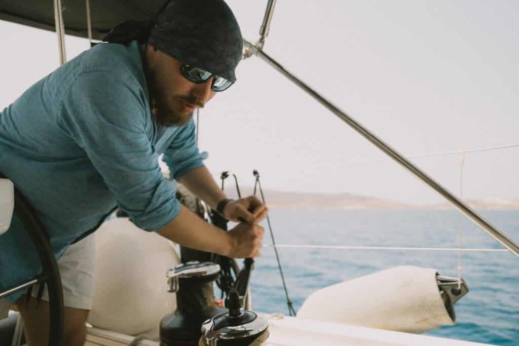 Ein Mann mit Kopftuch und Sonnenbrille bedient an einem sonnigen Tag die Winde eines Segelboots. Er trägt ein hellblaues Langarmhemd und weiße Shorts und genießt das Segeln, während hinter ihm das ruhige blaue Wasser glitzert und er in der Ferne die Küste sieht.