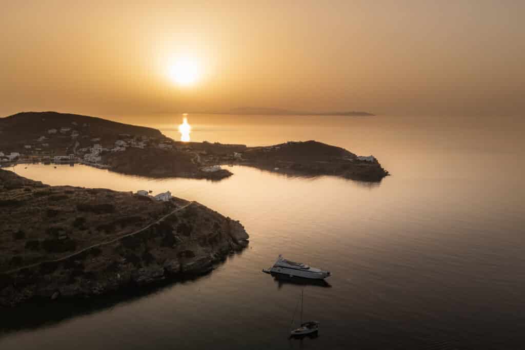 Ein heiterer Sonnenuntergang über einem ruhigen Meer, mit zwei Booten, die in der Nähe felsiger Ufer vor Anker liegen. Die reflektierte Sonne erzeugt einen goldenen Pfad auf dem Wasser, ideal für ein abendliches Segeln, während die Küste ein paar weiße Gebäude und schroffe Hügel im Hintergrund beherbergt. Der Himmel ist in warmen Orangetönen gemalt.