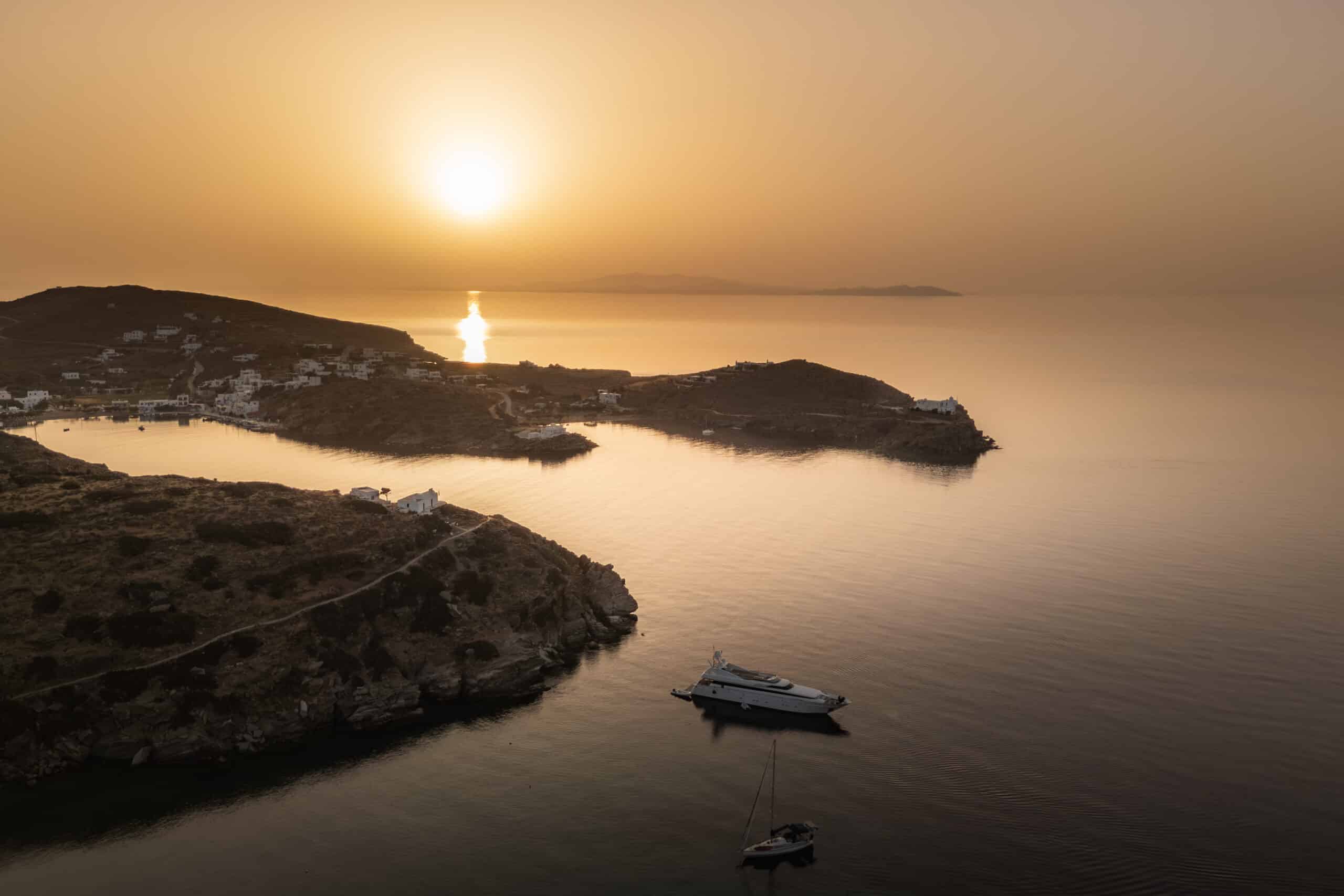 Ein heiterer Sonnenuntergang über einem ruhigen Meer, mit zwei Booten, die in der Nähe felsiger Ufer vor Anker liegen. Die reflektierte Sonne erzeugt einen goldenen Pfad auf dem Wasser, ideal für ein abendliches Segeln, während die Küste ein paar weiße Gebäude und schroffe Hügel im Hintergrund beherbergt. Der Himmel ist in warmen Orangetönen gemalt.