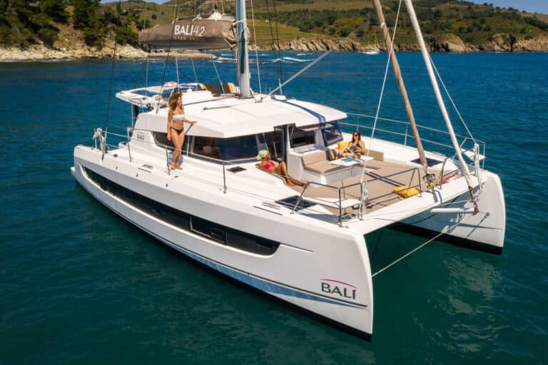 Ein weißer Bali 4.2 Segelkatamaran schwimmt auf klarem, blauem Wasser. Drei Personen entspannen sich auf dem Deck – eine liegt auf einer Sonnenliege, eine steht am Bug und eine andere liegt mit einem Buch zurückgelehnt da. Im Hintergrund sind grüne Hügel und Bäume unter einem blauen Himmel zu sehen, perfekt für einen Youngline Katamaran Segeltörn.