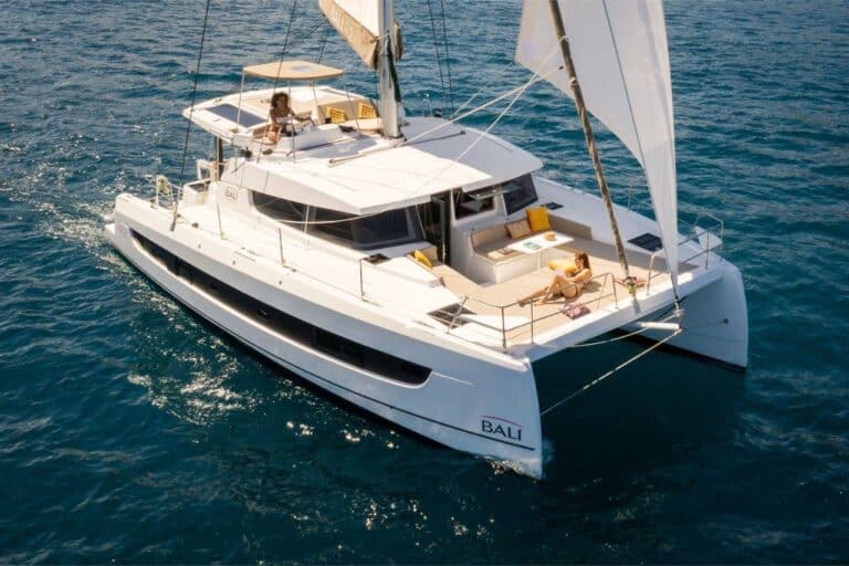 Ein weißer Youngline-Katamaran mit der Aufschrift „BALI“ auf den Seitensegeln auf klarem, blauem Wasser. Drei Personen entspannen auf dem Boot; eine auf dem Oberdeck und zwei auf dem Hauptdeck. Das Boot, perfekt für einen Segeltörn, hat Sonnenliegen und einen offenen Sitzbereich vorne.