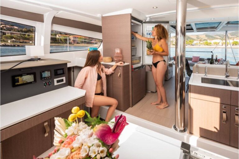 Zwei Frauen in Badeanzügen stehen in der Küche eines modernen Youngline Katamarans. Eine steht und legt eine Ananas in einen Schrank, während die andere sitzt und nach einem Snack greift. Es gibt Bedienfelder, Blumen auf der Theke und durch die Fenster blickt man auf das Meer und die Berge.