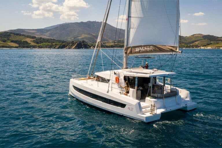 Ein schnittiger weißer Katamaran namens „Bali 4.2“ segelt auf ruhigen blauen Gewässern und nimmt Kurs auf einen malerischen Segeltörn. Das Youngline-Schiff verfügt über ein offenes Deck mit Sitzgelegenheiten, umgeben von atemberaubenden grünen Hügeln und fernen Bergen unter einem klaren, sonnigen Himmel mit vereinzelten Wolken.