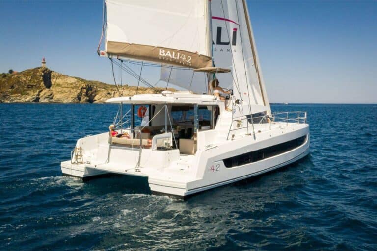 Ein weißer Youngline-Katamaran namens BALI 4.2 segelt in klarem, blauem Wasser in der Nähe von felsigem Küstengelände mit einem Leuchtturm auf einer Klippe. Das Boot hat ein großes Segel und mehrere Passagiere genießen das offene Deck unter dem sonnigen Himmel und sorgen für einen unvergesslichen Segeltörn.