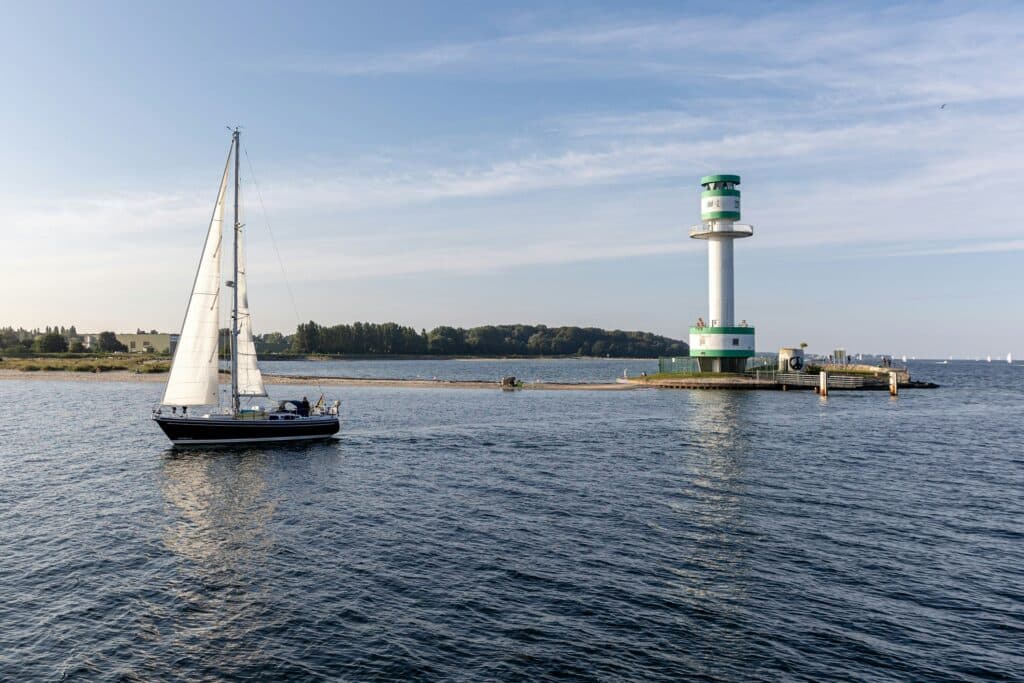Ostsee ist zu sehen und ein Segelboot. Das Wasser und der Himmel sind blau. Ein kleiner Leuchtturm ist auch zu sehen.