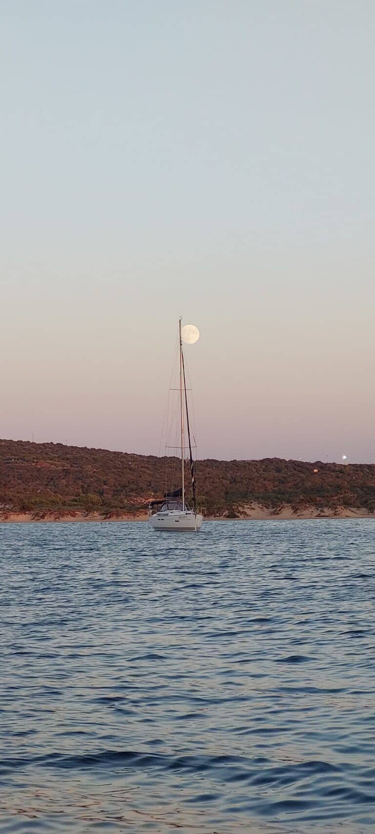 Ein Segelboot vor Anker in ruhigen Gewässern mit felsigen Hügeln im Hintergrund, das die ruhige Atmosphäre eines Segelurlaubs auf Sardinien einfängt. Der Vollmond steht tief am Himmel und erweckt den Eindruck, als säße er auf dem Mast des Segelboots. Der Himmel ist in sanften Farbtönen gehalten, die entweder Sonnenaufgang oder Sonnenuntergang anzeigen.