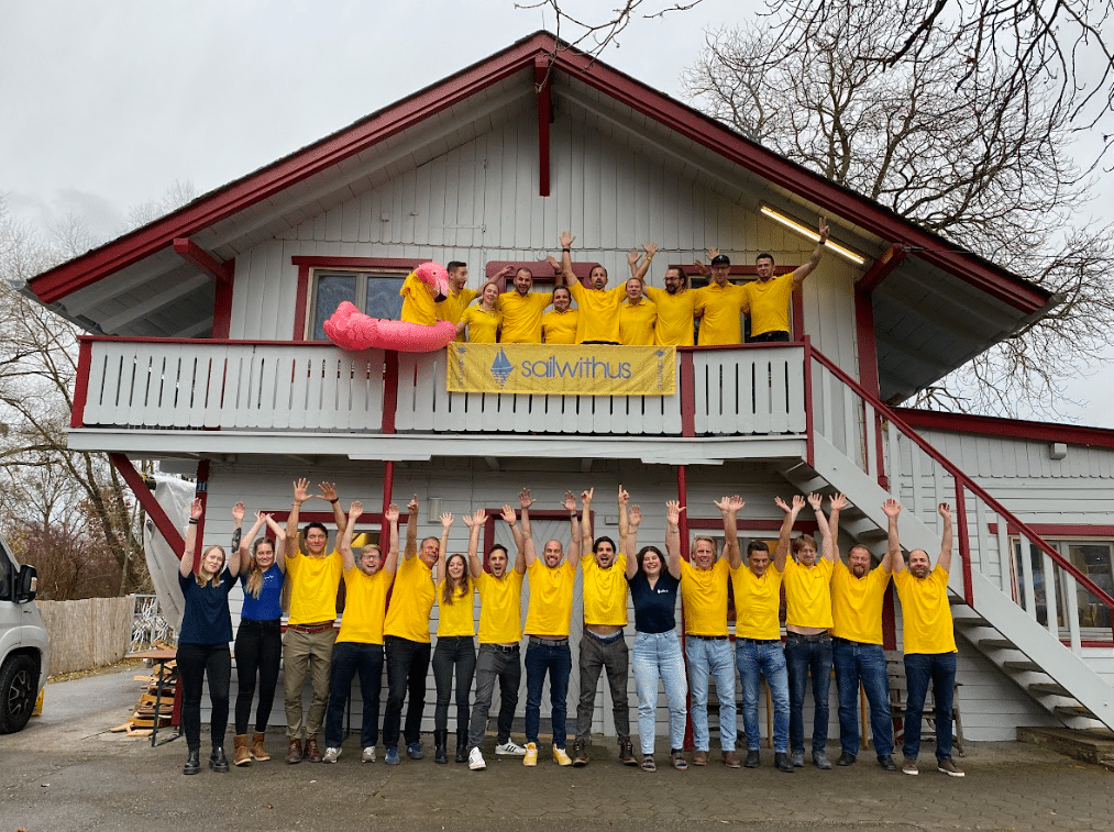 Eine Gruppe von Menschen in gelben Hemden steht vor einem zweistöckigen Holzgebäude, einige auf einem Balkon und andere auf dem Boden darunter, und alle strecken ihre Hände in die Luft. Auf einem Banner auf dem Balkon steht „Segel mit uns“. Auf dem Balkongeländer thront ein rosa Flamingo-Wagen. Es ist eine ihrer lebhaftesten Veranstaltungen!