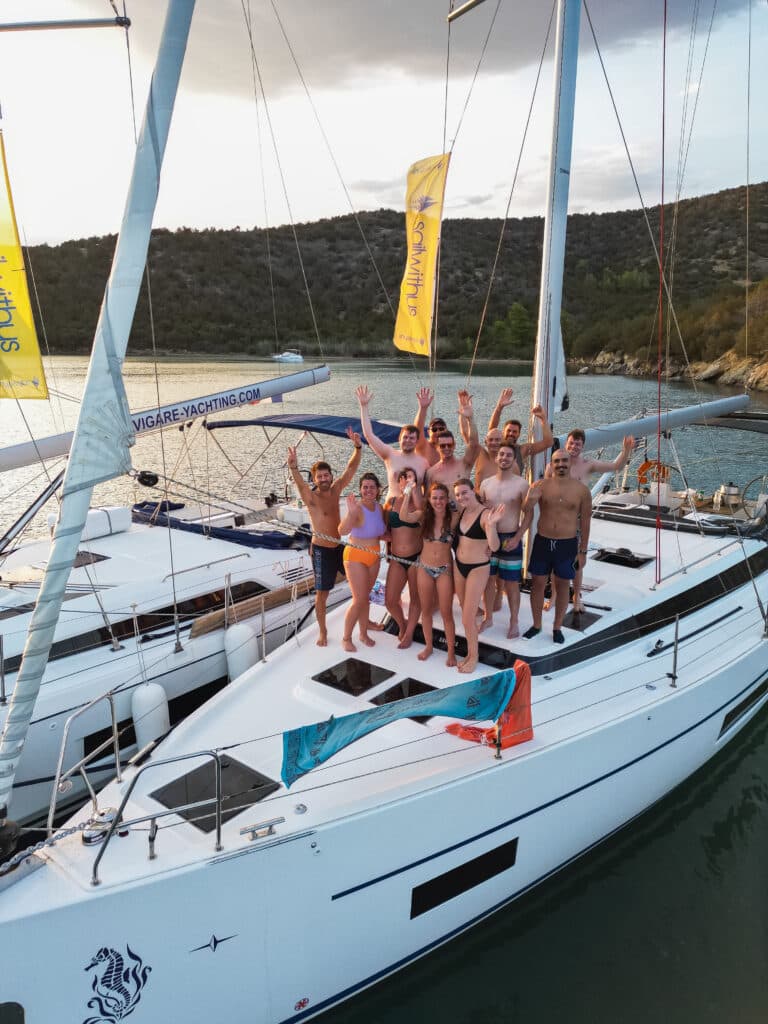 Eine Gruppe von Menschen steht lächelnd und winkend auf dem Deck einer weißen Yacht. Sie tragen Badebekleidung und scheinen einen sonnigen Tag während ihres Segelurlaubs auf Sardinien zu genießen. Neben ihnen ist eine weitere Yacht zu sehen, im Hintergrund sind malerische Hügel und Bäume zu sehen.