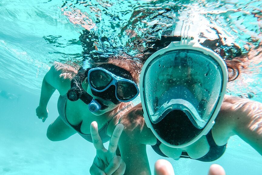 Zwei Personen unter Wasser mit Schnorchelmasken machen während ihres Segelurlaubs ein Selfie. Eine Person hält mit den Fingern ein Peace-Zeichen hoch. Sie sind umgeben von klarem blauem Wasser in der Nähe ihres Katamarans. Tipps für Reiseleiter: So gestalten Sie unvergessliche Gruppenreisen