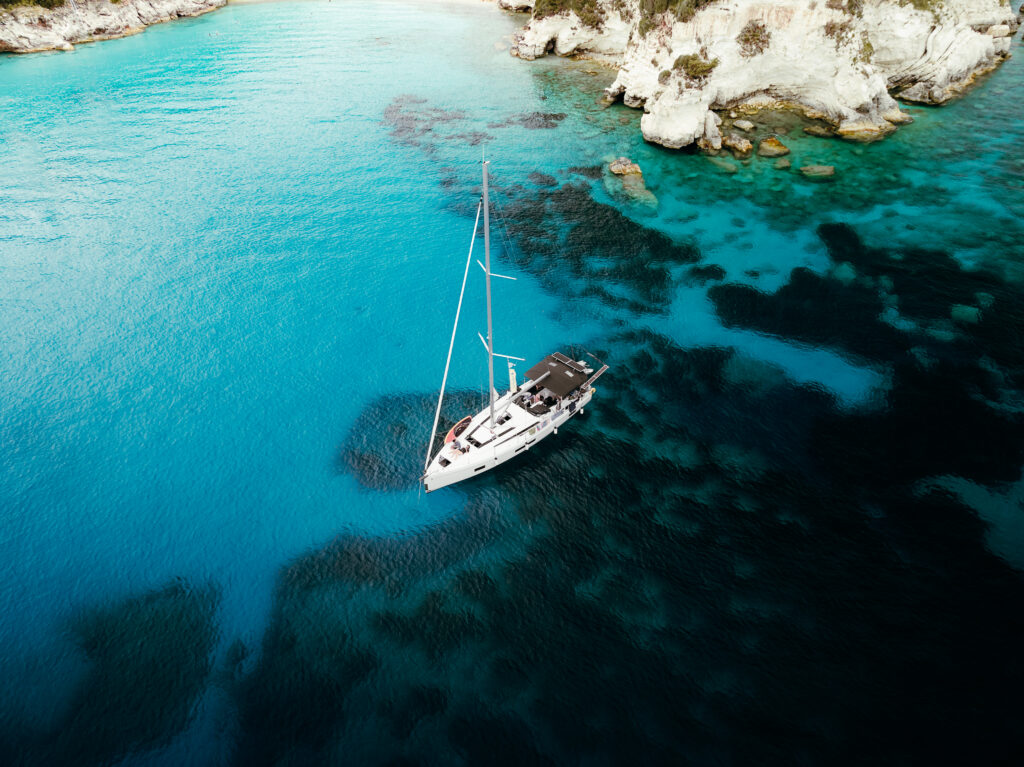 Ein Segelboot ankert in einer ruhigen, türkisfarbenen Bucht, umgeben von teilweise unter Wasser liegenden Felsen und weißen Klippen – perfekt für eine ruhige Pause während einer Atlantiküberquerung. Das Wasser variiert in Blautönen, mit Flecken von dunklerem Blau und Grün um das Boot herum.