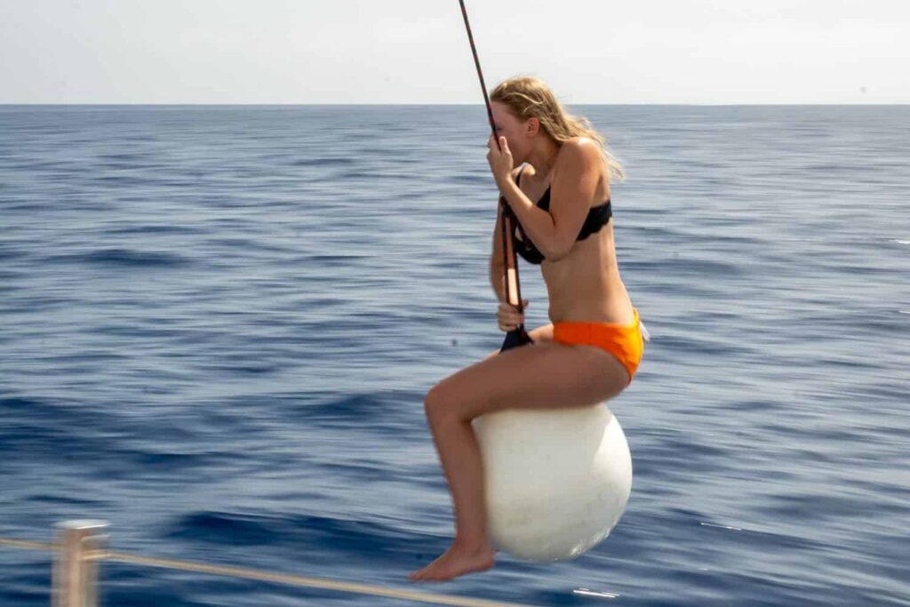 Eine junge Frau im orangefarbenen Bikini sitzt auf einer an einer Stange befestigten weißen Boje über dem tiefblauen Meer und genießt die individuelle Segelreise.