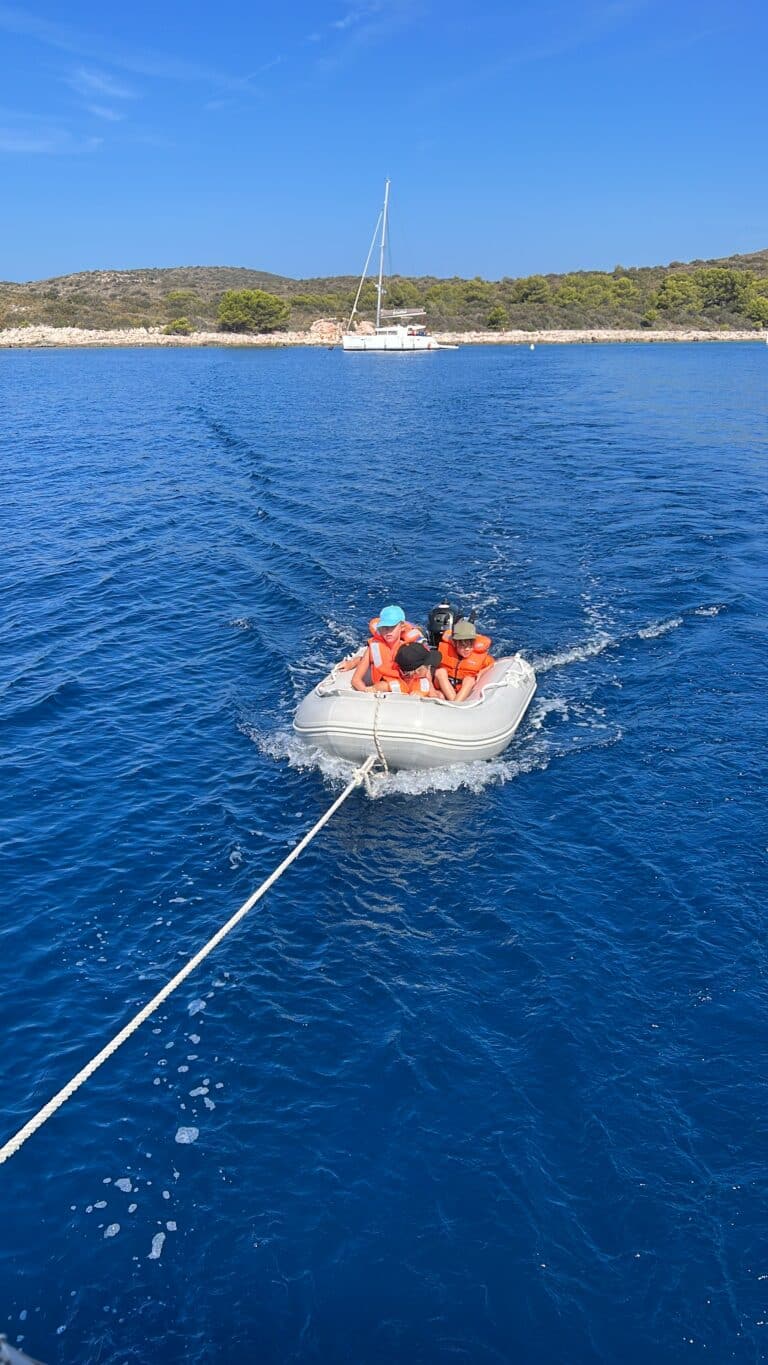 Eine Gruppe von Menschen mit Schwimmwesten fährt in einem kleinen Schlauchboot, das an einem Seil durch das blaue Meer gezogen wird. Im Hintergrund ankert ein Segelboot in der Nähe einer felsigen Küste mit grünen Bäumen unter einem klaren blauen Himmel und zeigt den Charme des familienfreundlichen Segeltörns im Mittelmeer.